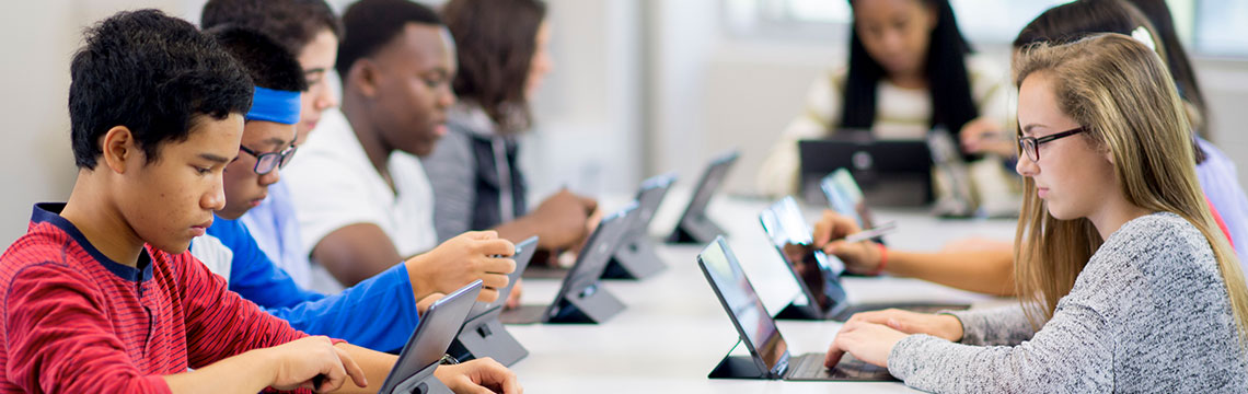 Students sitting at a large table working on laptop computers and tablet devices.