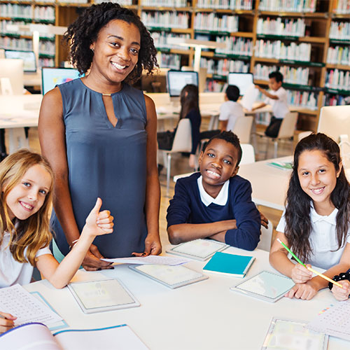 An elementary school teacher standing with three students seated at a table in the school library